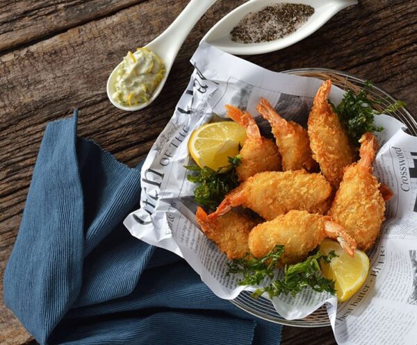 A basket of fried prawn with lemon wedges and sauce on a newspaper, on a wooden table